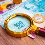 o que é inflação - Cédulas de real brasileiro com uma lupa sobre elas e algumas moedas apoiadas em um móvel de madeira