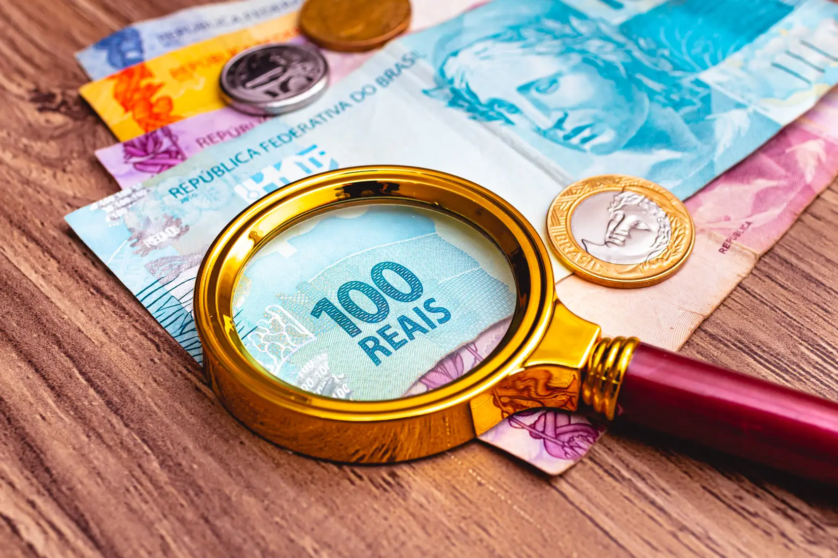 o que é inflação - Cédulas de real brasileiro com uma lupa sobre elas e algumas moedas apoiadas em um móvel de madeira