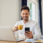 o que é previdência privada - homem sorrindo em frente ao notebook, olhando para o celular e tomando café
