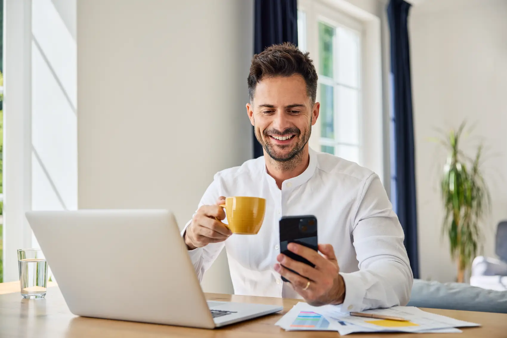 o que é previdência privada - homem sorrindo em frente ao notebook, olhando para o celular e tomando café