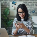 tendencias-do-e-commerce mulher navegando no celular sorrindo com um notebook aberto na mesa de escritório