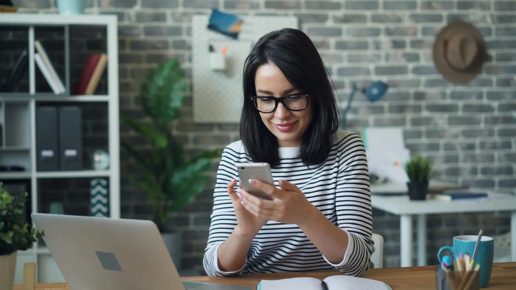 tendencias-do-e-commerce mulher navegando no celular sorrindo com um notebook aberto na mesa de escritório