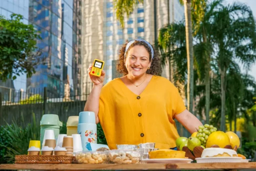 Dia do Empreendedorismo Feminino - mulher branca de cabelos cacheados em frente à banca de café da manhã na rua, com maquininha pagbank na mão