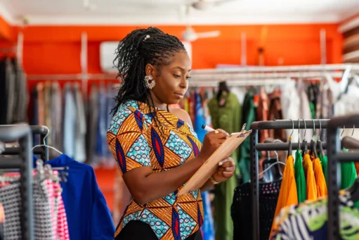 Obrigações do MEI - empresária de moda verificando o inventário em sua loja de roupas com sua prancheta