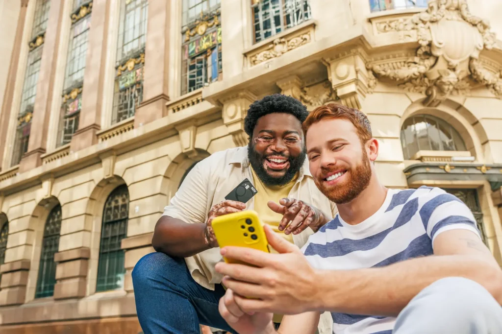 pagbank é seguro - homem negro com vitiligo e homem branco olham o celular, sorridentes, em frente a um prédio histórico. Homem negro segura cartão PagBank.