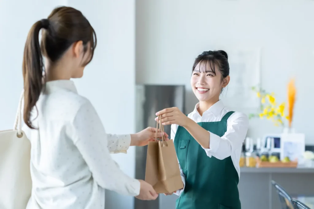 dia do cliente 2026 - Uma jovem japonesa trabalhando como garçonete em um café, entregando uma sacola À cliente