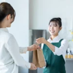 dia do cliente 2026 - Uma jovem japonesa trabalhando como garçonete em um café, entregando uma sacola À cliente