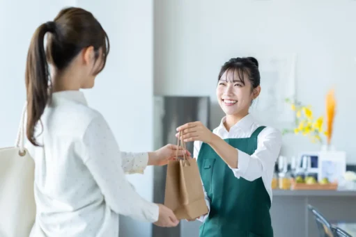 dia do cliente 2026 - Uma jovem japonesa trabalhando como garçonete em um café, entregando uma sacola À cliente