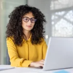 como-cobrar-um-cliente- Uma mulher de cabelo encaracolado, usando óculos e camisa amarela, trabalha em um laptop em um ambiente de escritório.
