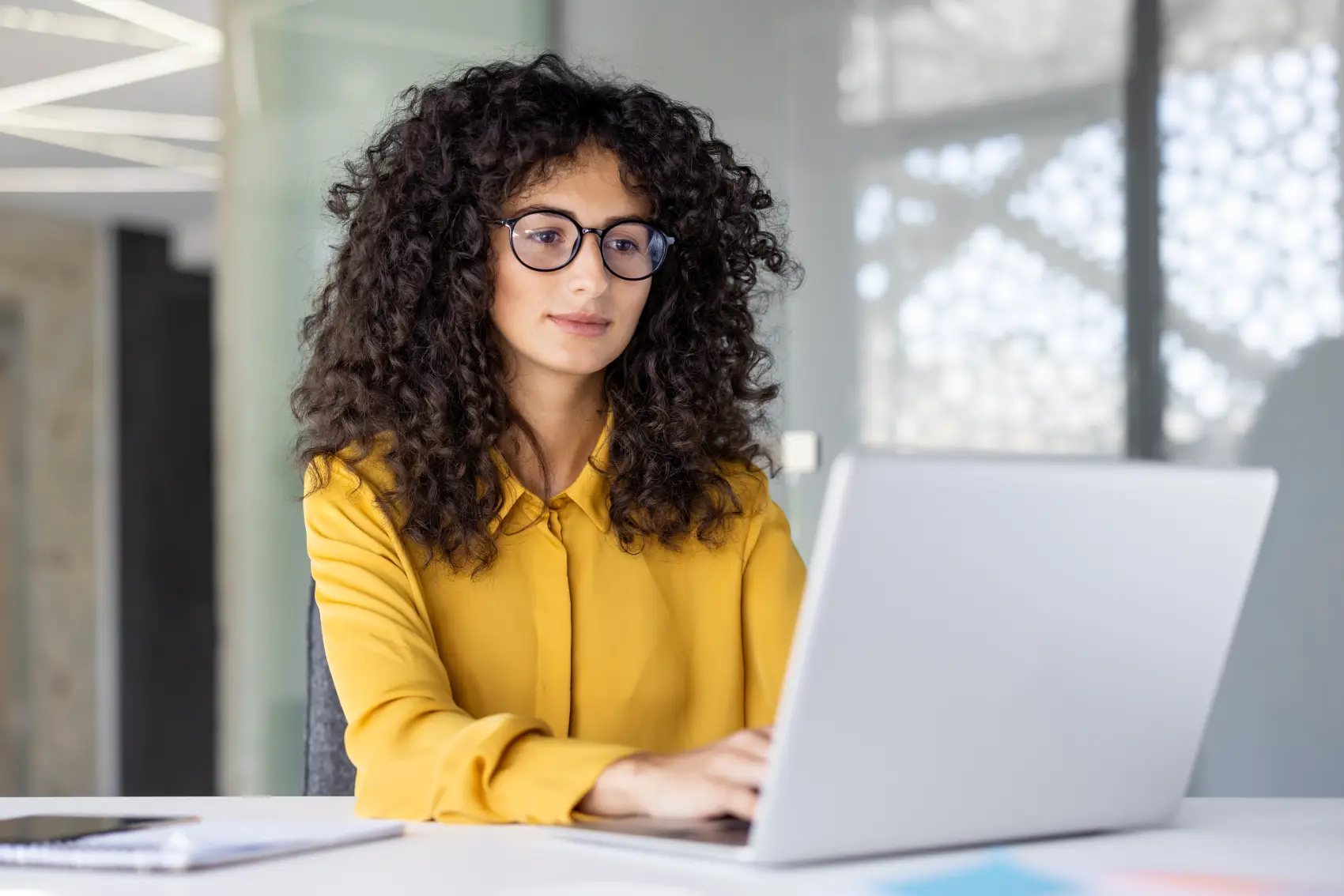 como-cobrar-um-cliente- Uma mulher de cabelo encaracolado, usando óculos e camisa amarela, trabalha em um laptop em um ambiente de escritório.