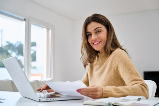 como cobrar um cliente - Mulher de meia-idade feliz olhando para a câmera e segurando documentos enquanto trabalha em uma mesa em casa