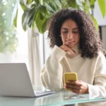 empréstimo para quitar dívidas - mulher negra feliz e pensativa em frente ao celular