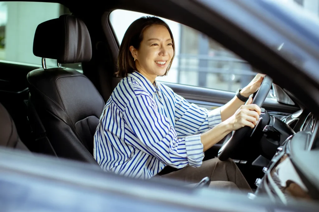 o-que-e-ipva Uma coreana senta-se no banco do motorista de um carro. Ela está sorrindo e focada na estrada à frente