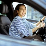 o-que-e-ipva Uma coreana senta-se no banco do motorista de um carro. Ela está sorrindo e focada na estrada à frente