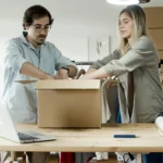como-montar-uma-loja-virtual homem e mulher ajustando uma caixa de papelão em uma mesa de escritório com várias caixas ao redor e um notebook sobre a mesa