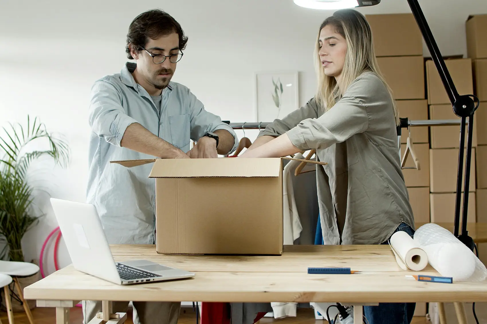 como-montar-uma-loja-virtual homem e mulher ajustando uma caixa de papelão em uma mesa de escritório com várias caixas ao redor e um notebook sobre a mesa