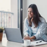 darf-e-imposto-de-renda Mulher positiva conversando com o cliente em uma chamada de vídeo no laptop, tomando notas, falando no computador, sentando na mesa de trabalho, usando dispositivos para comunicação de trabalho