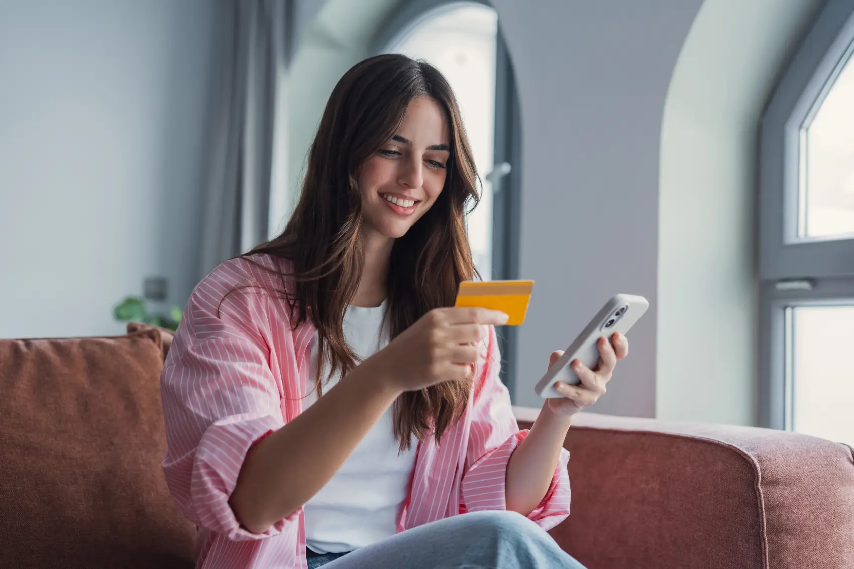 debito-e-credito-diferencas Uma mulher latina sorridente segura seu smartphone e cartão de crédito enquanto faz um pagamento on-line, desfrutando de serviços bancários móveis seguros e transações digitais rápidas no conforto de sua casa