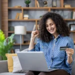 frete grátis​ - Jovem mulher triunfante comprando mercadorias on-line com um cartão de crédito, usando um laptop, feliz sentado em um sofá em sua sala de estar, celebrando uma transação bem-sucedida