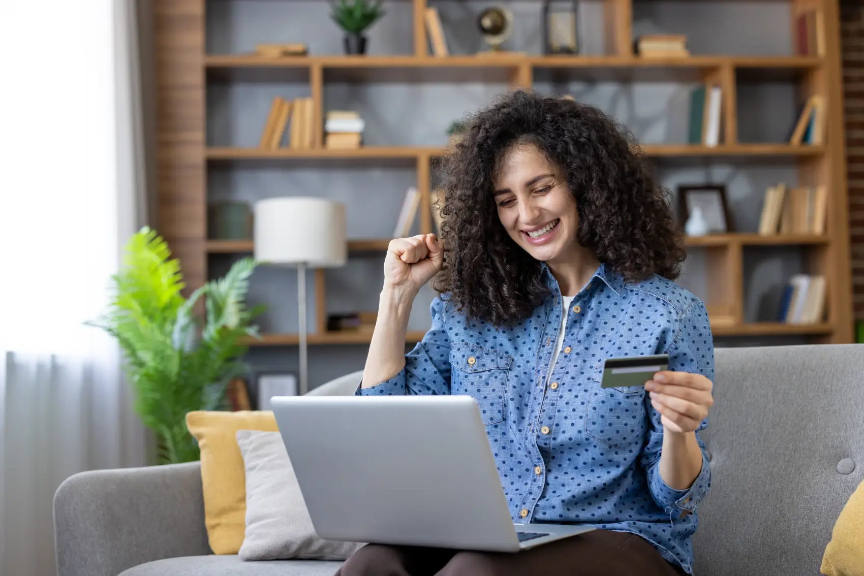 frete grátis​ - Jovem mulher triunfante comprando mercadorias on-line com um cartão de crédito, usando um laptop, feliz sentado em um sofá em sua sala de estar, celebrando uma transação bem-sucedida