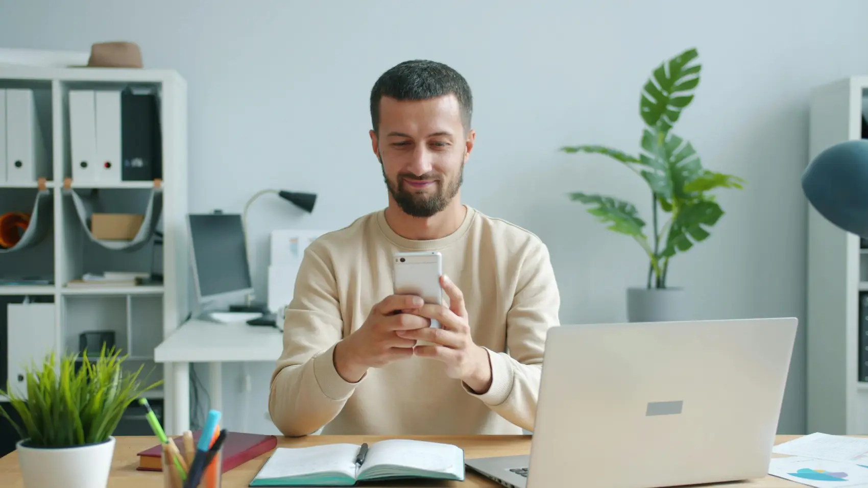 controle-de-pedidos homem com celular na mão em uma mesa de escritório com caderno e notebook abertos