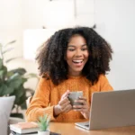 Lucro do FGTS - Mulher negra feliz desfrutando café da manhã enquanto usa laptop em casa.