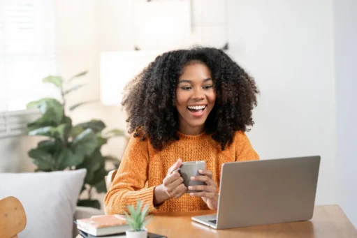 Lucro do FGTS - Mulher negra feliz desfrutando café da manhã enquanto usa laptop em casa.