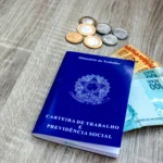 décimo terceiro salário - carteira de trabalho em mesa com notas de cem e cinqunta reais e algumas moedas de real