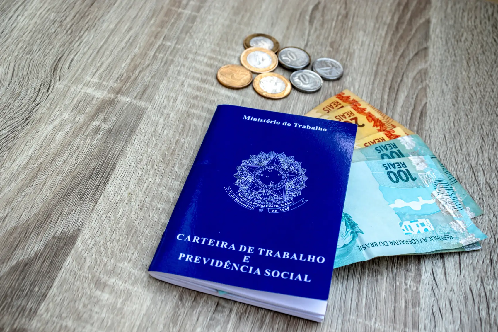 décimo terceiro salário - carteira de trabalho em mesa com notas de cem e cinqunta reais e algumas moedas de real