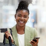 como funciona o parcelamento no cartão de crédito​ - mulher negra segurando cartão de crédito e sacola de compras em uma mão e celular na outra. ela sorri para a câmera com os cabelos presos em um coque alto