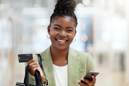 como funciona o parcelamento no cartão de crédito​ - mulher negra segurando cartão de crédito e sacola de compras em uma mão e celular na outra. ela sorri para a câmera com os cabelos presos em um coque alto
