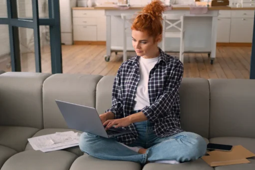 o que é amortização de empréstimo - Mulher caucasiana sentada no sofá de casa usando laptop