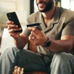 pix crédito -homem sentado no sofá olhando para o smartfone e cartão de crédito, sorrindoi