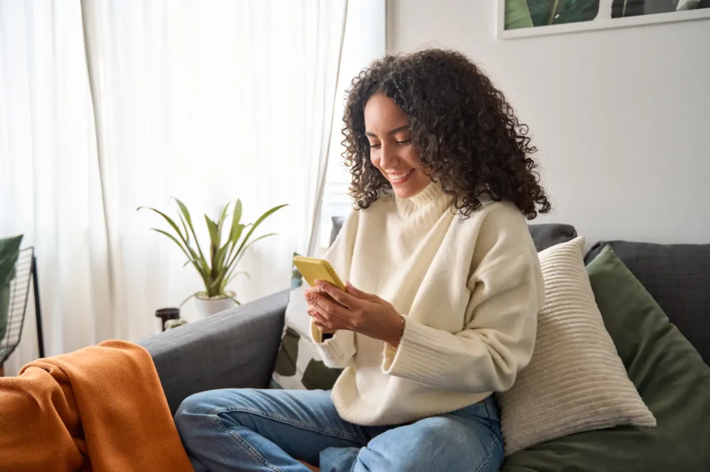 pix parcelado​ - Mulher jovem e feliz, latina, sentada no sofá, usando o celular para fazer compras online, enviar mensagens e conversar confortavelmente na sala de estar de casa.