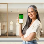 curso de trading - mulher mostrando app do celular em frente à sua loja de roupas