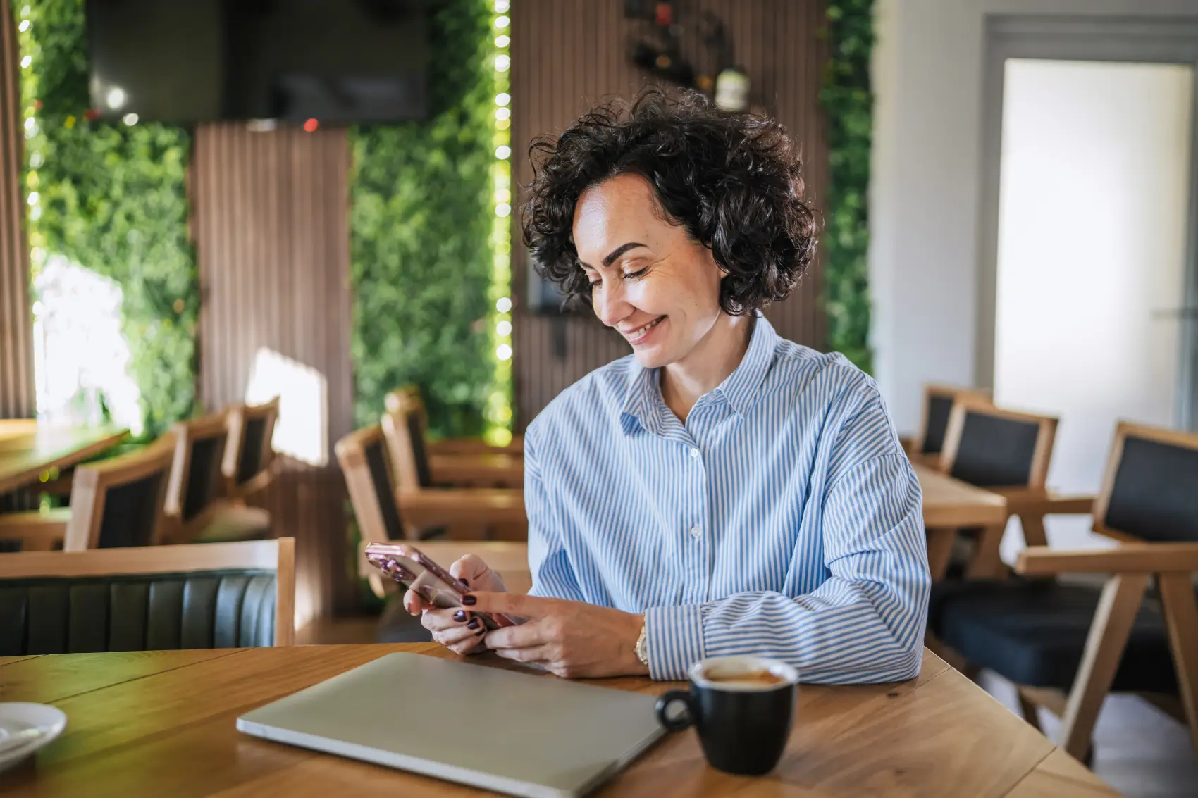 clt pode ser mei​ - Mulher com cabelo encaracolado sentada em uma mesa de madeira em um café moderno, verificando seu smartphone enquanto toma café expresso, laptop perto contra uma parede de planta verde,