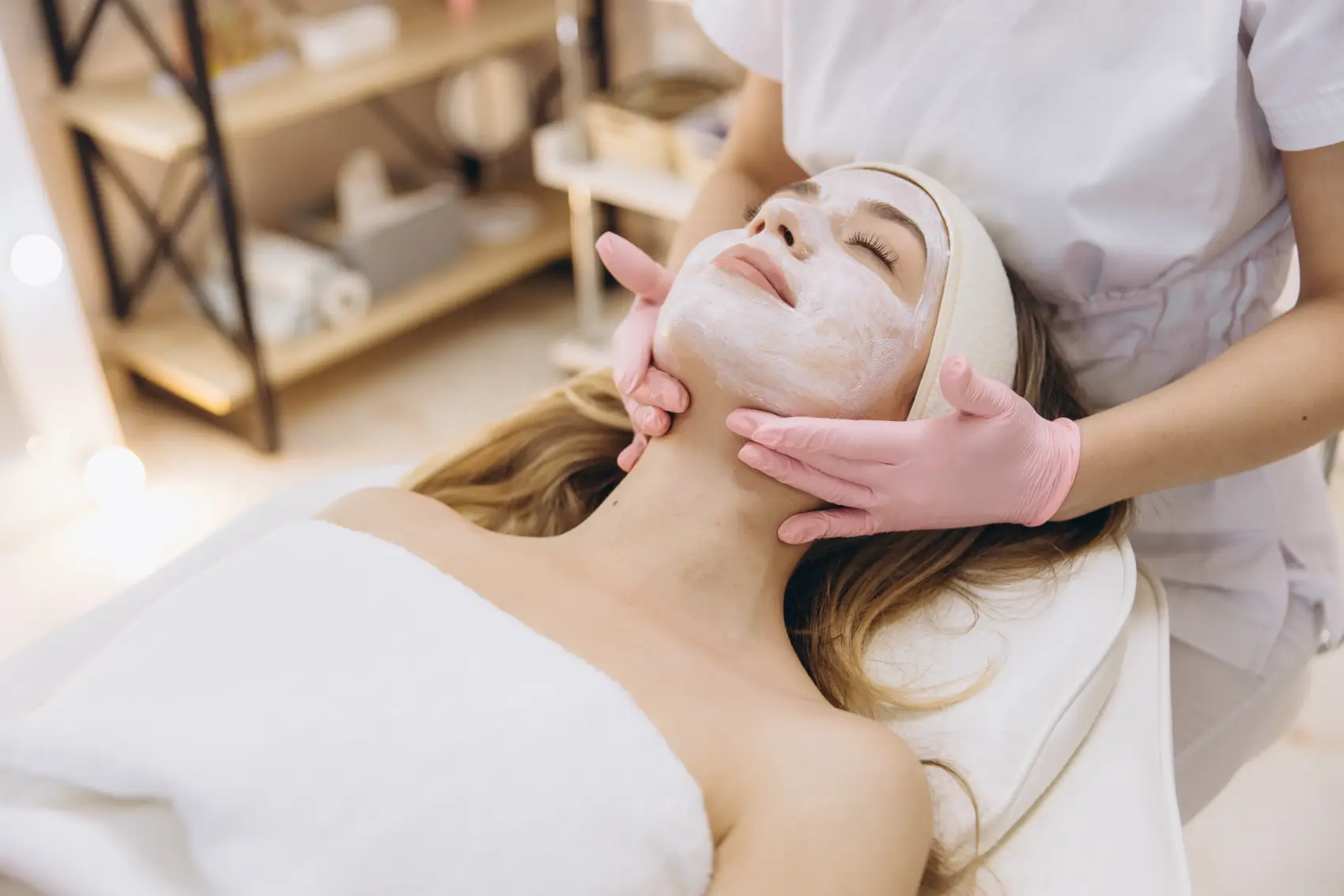 Esteticista pode ser MEI? - Esteticista vestindo luvas rosa aplicando uma máscara facial hidratante a uma mulher relaxada deitada em uma mesa de massagem em um salão de beleza