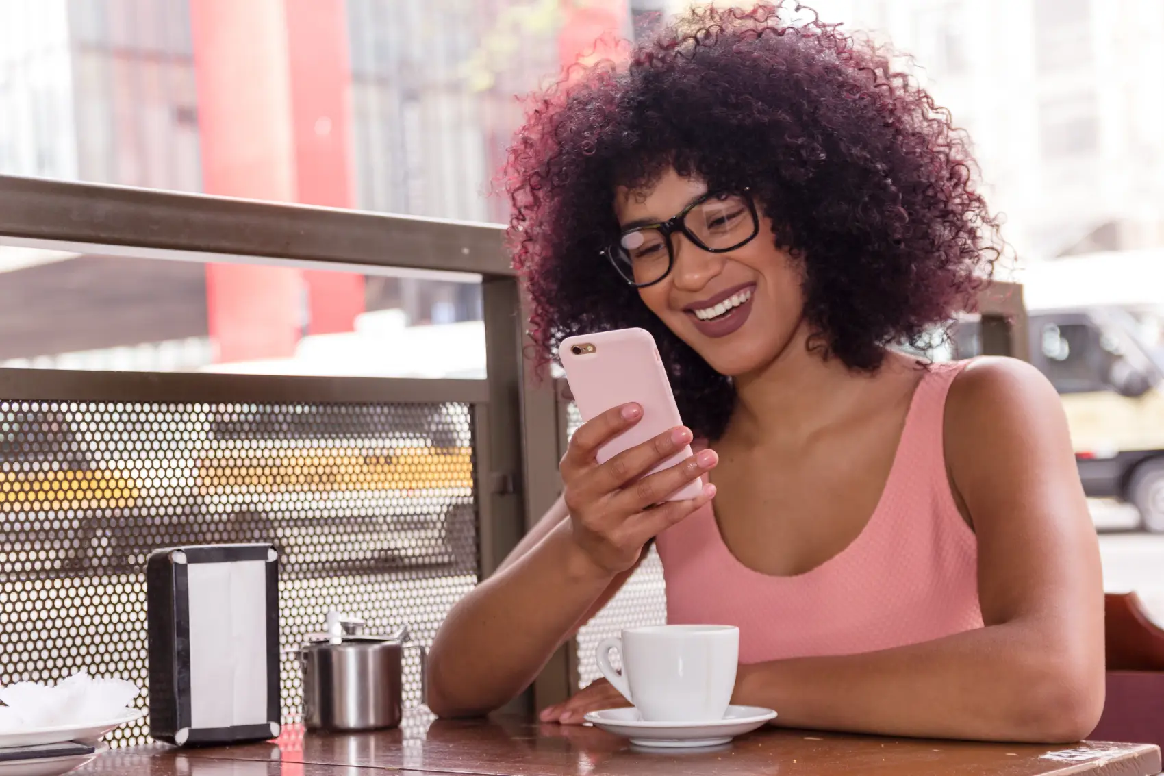 PagBank Premiado - Linda mulher negra de óculos usando tecnologia ao ar livre e tomando café na cidade de São Paulo durante o verão, com um cenário urbano ao fundo