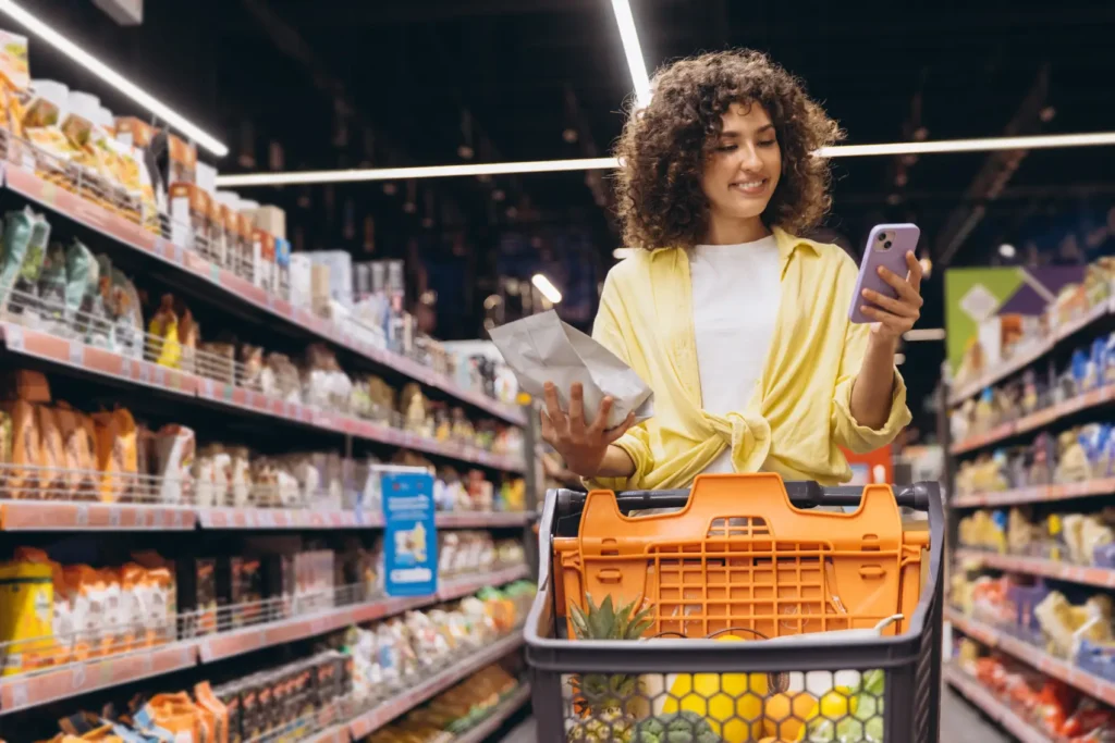 como-precificar-um-produto Jovem mulher comparando produto na mão com seu aplicativo de smartphone, encontrando melhores ofertas e gerenciando suas compras de supermercado