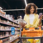 como-precificar-um-produto Jovem mulher comparando produto na mão com seu aplicativo de smartphone, encontrando melhores ofertas e gerenciando suas compras de supermercado