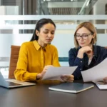 simples nacional 2027 - Duas empresárias diversas revisando documentos e discutindo trabalho, colaborando em um projeto ou relatório enquanto sentadas em uma mesa em um ambiente de escritório corporativo moderno