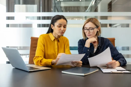 simples nacional 2027 - Duas empresárias diversas revisando documentos e discutindo trabalho, colaborando em um projeto ou relatório enquanto sentadas em uma mesa em um ambiente de escritório corporativo moderno
