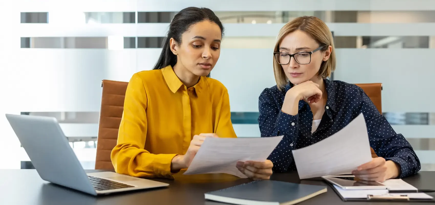 simples nacional 2027 - Duas empresárias diversas revisando documentos e discutindo trabalho, colaborando em um projeto ou relatório enquanto sentadas em uma mesa em um ambiente de escritório corporativo moderno
